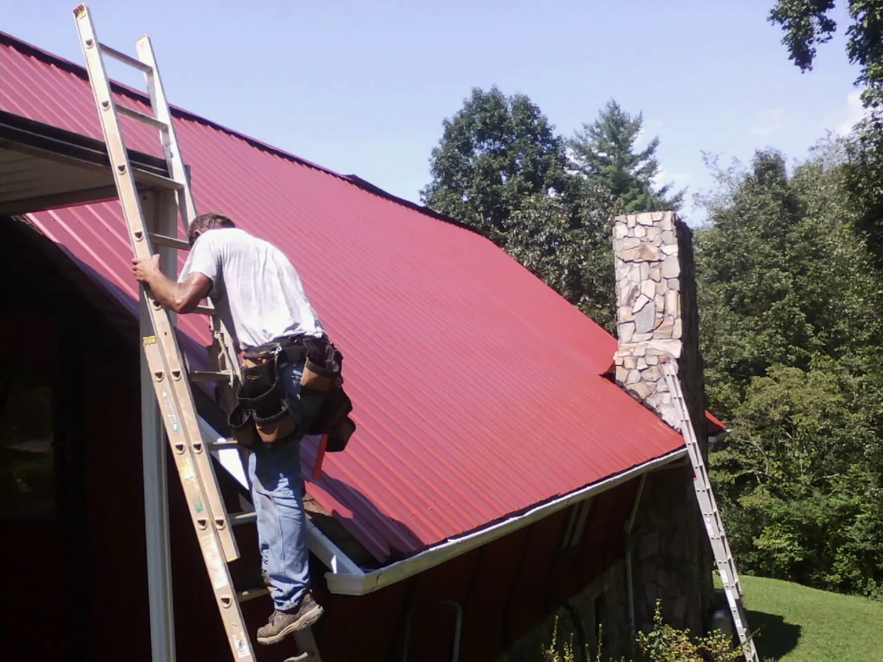 Roofer on a ladder beside a red metal roof and stone chimney — Soffit & Fascia Repair by Roofing Brothers in Jeddo